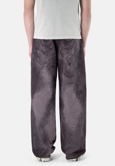 Person trägt dunkelgraue Jeans mit weitem Bein und Gesäßtaschen sowie ein hellgraues T-Shirt und steht vor einem einfarbigen Hintergrund.