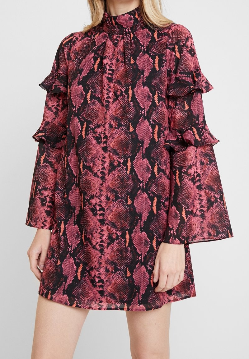 Femme portant une robe rose et noire à imprimé serpent avec de longues manches cloche et des détails à volants sur le haut des bras et un col haut.