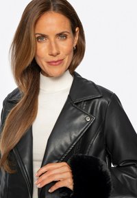 Schwarze Kunstlederjacke mit Reverskragen und Fellmanschetten, getragen über einem weißen gerippten Rollkragenpullover. Das Model hat lange braune Haare.