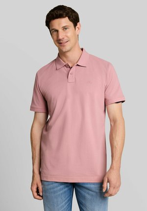 Man draagt een lichtroze poloshirt met korte mouwen en blauwe spijkerbroek, staand tegen een effen grijze achtergrond.