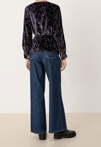 Top in velluto con un motivo viola e nero, affusolato in vita, abbinato a jeans a gamba larga blu e stivaletti neri.