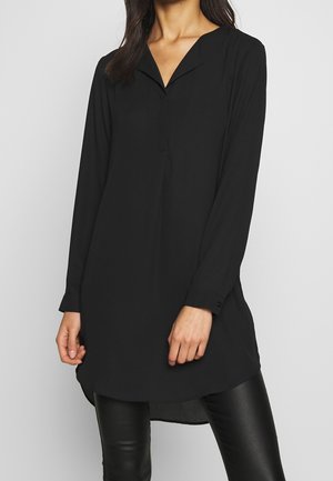 Frau trägt eine schwarze Tunika-Bluse mit langen Ärmeln und Kragen sowie schwarze Lederhose, steht vor einem einfarbigen grauen Hintergrund.
