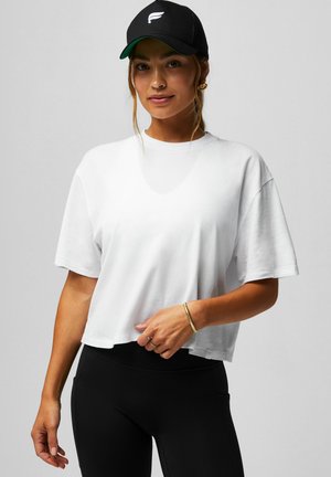 Frau mit schwarzer Mütze, weißem locker sitzendem T-Shirt, schwarzen Leggings, goldenen Creolen, Armband und Ringen, steht vor einem schlichten grauen Hintergrund.