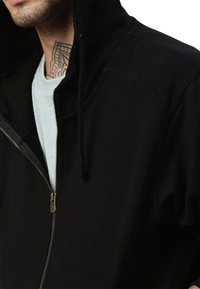 Giacca nera con cappuccio e zip, dal tessuto liscio. Presenta una zip argentata e cuciture a vista. Abbinata a una maglia grigia chiara con collo rotondo sotto.