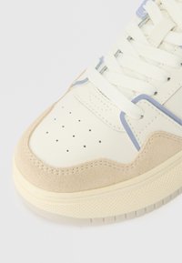 Lenkkari, jossa on kerma väri nahka päällinen, vaalean beige mokkanahka korostuksia, reikiä varpaassa, sinisiä yksityiskohtia ja tekstuurinen kumipohja.