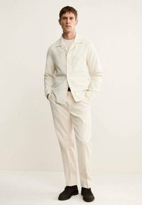 Blazer de color crema con un bolsillo, combinado con pantalones a juego. El conjunto presenta un diseño limpio y una textura sutil. Zapatos oscuros completan el look.