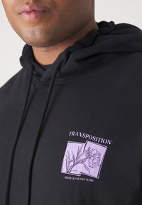 Nahaufnahme einer Person in einem schwarzen Hoodie mit einem lila Blumenmuster und dem Text "TRANSPOSITION" und "GROW WITH THE FLOW" auf der Brust.