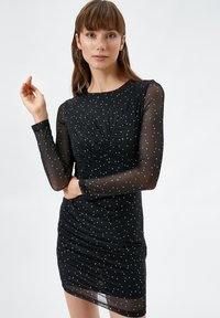 Zwarte jurk met lange doorzichtige mouwen, versierd met witte stippen. Heeft een gerimpeld ontwerp en een getailleerde silhouet, met een enkelhoge snit.