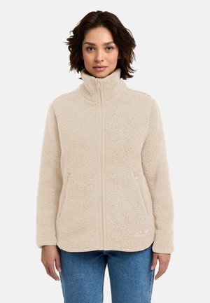 Vrouw draagt een beige fleecejas met rits en hoge kraag en een blauwe spijkerbroek, staand tegen een effen lichte achtergrond.