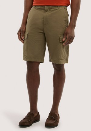 Man draagt olijfgroene cargoshorts met klepzakken, bruine loafers en een rood shirt tegen een eenvoudige lichte achtergrond.