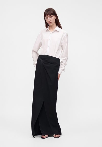 Femme aux longs cheveux bruns portant une chemise blanche boutonnée et une longue jupe portefeuille noire avec des sandales, debout devant un fond uni.
