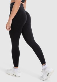 Leggings neri con vita alta, design aderente, texture liscia e branding minimale, abbinati a scarpe sportive bianche e calzini.