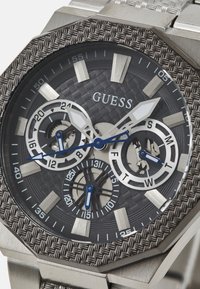 Reloj de acero inoxidable gris con caja octogonal, esfera texturizada y tres subesferas. Presenta detalles en azul y marcadores de hora luminosos.