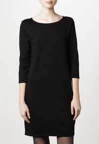 Robe noire en tissu doux, avec un col rond, des manches trois-quarts et une coupe ajustée, se terminant juste au-dessus du genou.