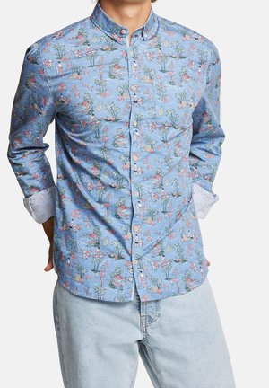 Mann trägt hellblaues Hemd mit buntem tropischem Muster und hellblaue Jeans, Hände hinter dem Rücken.