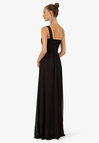 Sort maxi-kjole med smocket bodice, brede skulderstropper og flydende stof. Ryggen er åben, hvilket skaber en svajende silhuet.