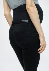 Čierne tehotenské džínsy s elastickým pásom a pútkami na opasok. Obsahujú logo nášivku z umelého kože a zadné vrecká. Hladká textúra.