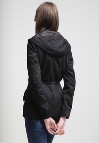 Czarna kurtka z kapturem i sznurkiem w talii, o gładkiej fakturze i prostym kroju. Widoczne przeszycia wzdłuż szwów.