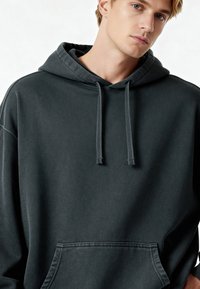 Sötét szürke kapucnis pulóver, laza fazon, állítható zsinórokkal és elülső kenguruzsebbel. Puha, texturált anyag.