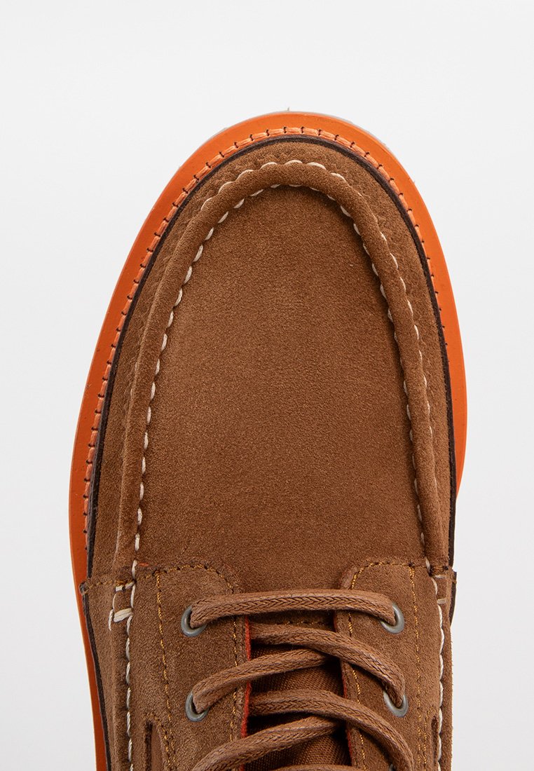 Braune Wildleder-Mokassins mit orangefarbener Gummisohle, erhöhten Nähten und Schnürdetails, die eine glatte Textur und eine abgerundete Zehenpartie betonen.