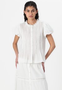 Hvit bluse med korte ermer, plisserte detaljer, blondekrage og knappestenging. Kombinert med en matchende hvit lagdelt skjørt.