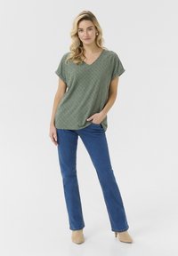 Green, perforated short-sleeve top with a V-neck, paired with blue jeans. The top features a loose fit and intersecting diamond pattern.
