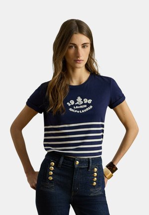 Žena nosi tmavomodré tričko s bielym logom kotvy a textom "1996 Lauren Ralph Lauren", doplnené tmavými džínsami s vysokým pásom a zlatými gombíkmi.