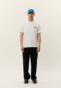 Krēmkrāsas T-krekls ar sarkanu "LD" logotipu, kombinēts ar melnām biksēm un brūnām kurpēm. Outfitu papildina zila cepure, kas nodrošina ikdienišķu izskatu.