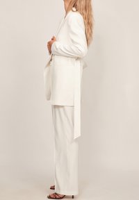 Blazer blanc ajusté avec une ceinture à nouer, présentant un col cranté et deux poches avant, associé à un pantalon large assorti.