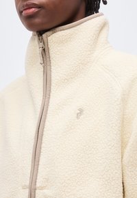 Κοντινό πλάνο ατόμου που φοράει ένα εκρού, υφασμάτινο φλις μπουφάν με ψηλό γιακά και μπεζ περιμετρικό φερμουάρ, εμφανίζοντας μερικώς το πρόσωπο και την περιοχή του στήθους.