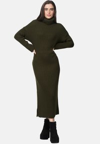 Olivgrünes, geripptes Strickkleid mit hohem Rollkragen, langen Ärmeln und seitlichen Schlitzen; kombiniert mit schwarzen Plateau-Stiefeletten.