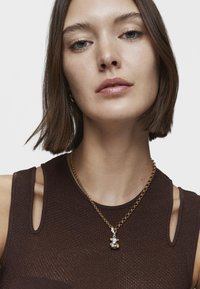 Collar de cadena de oro con charms de perlas irregulares. La modelo lleva una blusa sin mangas de color marrón con tejido texturizado y cortes en los hombros.