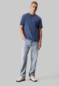 T-shirt in cotone blu con collo rotondo e un piccolo logo ricamato; abbinato a jeans in denim chiaro e scarpe nei toni neutri.