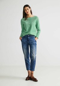 Grünes Langarmoberteil mit einem strukturierten Muster, kombiniert mit verblichenen blauen Denim-Jeans. Braune Socken und dunkle Slipper vervollständigen das Outfit.
