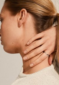 L'anello a onda in argento riposa su un dito, evidenziando un design liscio e minimalista. È visibile il tono della pelle e un maglione lavorato a maglia con texture.