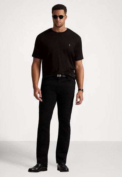 Svart bomulls-t-shirt med korta ärmar och rund hals, kombinerad med svarta jeans. Person som bär solglasögon och svarta skor, stående mot en vit bakgrund.