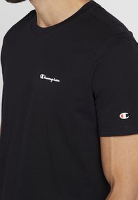 Black cotton t-shirt with a crew neckline. Features a white “Champion” logo on the chest and a small logo in red and white on the sleeve.