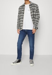 Grå och svart rutigt flanellskjorta över en vit t-shirt, tillsammans med blå jeans och svarta högstövlar med vita sulor.