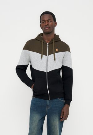 Moški v barvno razdeljenem puloverju z zadrgo v rjavi, sivi in črni barvi ter modrih kavbojkah, stoji z eno roko v žepu.