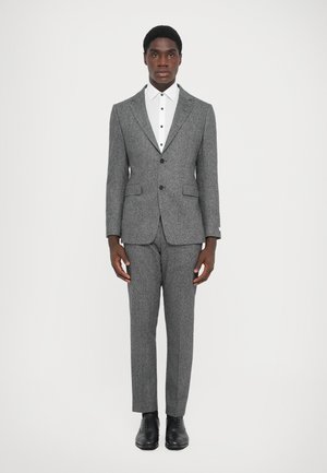 Fato cinza com padrão tipo tweed e lapela recortada, acompanhado por uma camisa branca com botões pretos. Combinado com sapatos de vestir pretos.