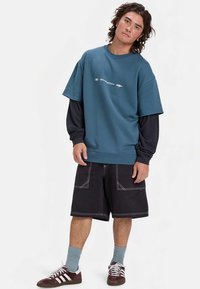 Kék, túlméretezett pulóver kerek nyakkal, fehér grafikával, fekete hosszú ujjú póló fölött viselve. Fekete rövidnadrággal, kék zoknival és sportcipővel párosítva.