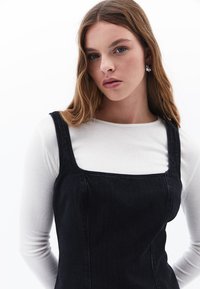 Schwarzes Denim-Kleid mit quadratischem Ausschnitt und breiten Trägern, mit vertikalen Stickdetails, getragen über einem weißen Langarmshirt.