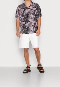 Różowa koszula z krótkim rękawem w czarny wzór liści, zestawiona z białymi szortami i czarnymi sandałami. Projekt ma luźny krój i zapięcie na guziki.