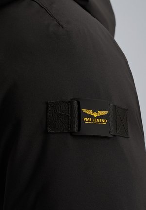 Svart vattentät jacka med en rektangulär märkeslapp med gul text och logotyp, tillsammans med texturerade svarta remmar.
