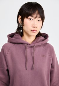 Lilla hettegenser med kenguru-lomme foran, justerbare snorer og en liten logo på brystet. Stoffet ser mykt og teksturert ut.