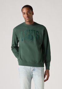 Zelená mikina so vzorovaným logom "LEVI'S" na hrudi, okrúhly výstrih, rebrované manžety a spodný lem. Kombinovaná so svetlomodrými džínsami.