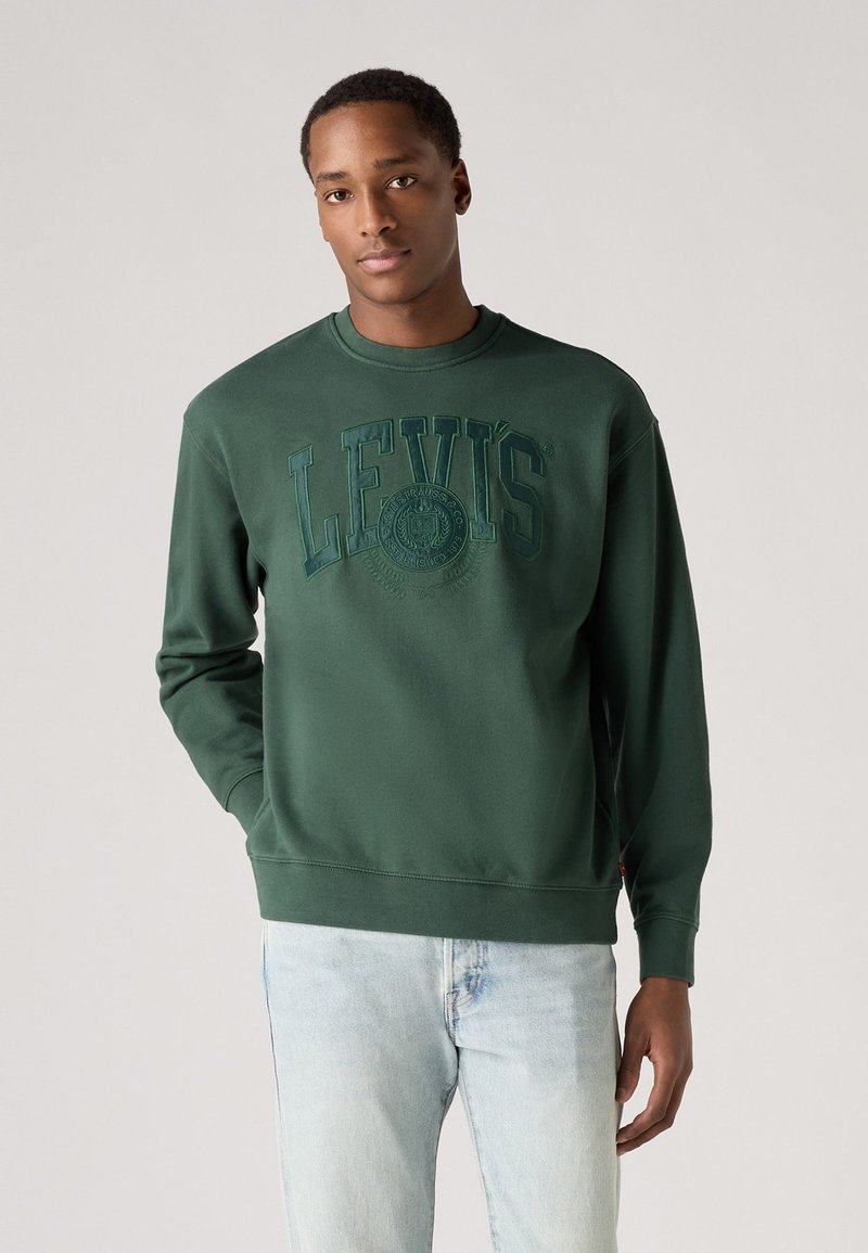 Zelená mikina so vzorovaným logom "LEVI'S" na hrudi, okrúhly výstrih, rebrované manžety a spodný lem. Kombinovaná so svetlomodrými džínsami.