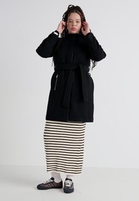 Svart zip-up jakke med belte, med lange ermer og strukturert overflate. Kombinert med et striped maxi skjørt og joggesko.