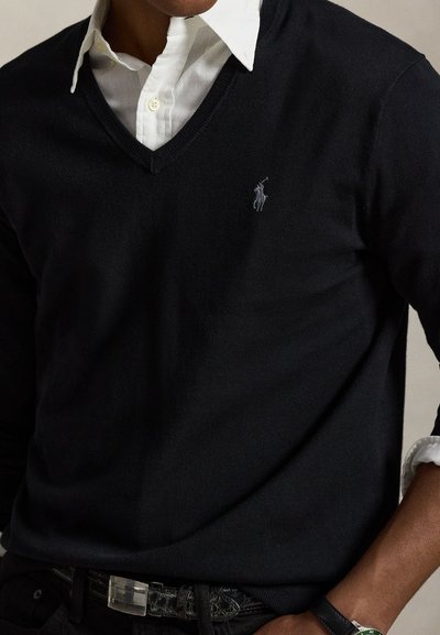 Mann trägt ein weißes Hemd mit Kragen unter einem schwarzen V-Ausschnitt-Pullover mit kleinem Polo-Spieler-Logo auf der Brust.