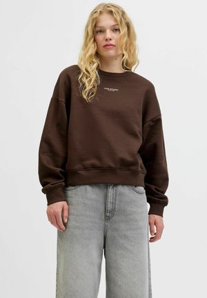 Brauner, übergroßer Pullover mit Rundhalsausschnitt und gerippten Bündchen; zeigt das weiße Textlogo "JJXX STUDIO" auf der Vorderseite. Getragen mit grauen Jeans.